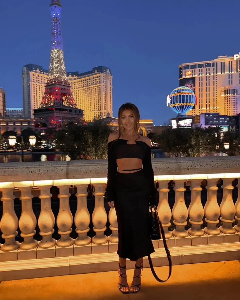 Woman in a stylish black outfit poses near the illuminated Eiffel Tower replica in Las Vegas at night.