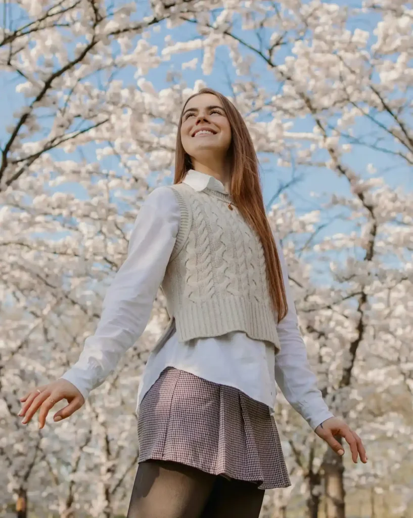 Young woman in a cable knit vest smiles joyfully amidst blooming cherry blossom trees under a clear blue sky.