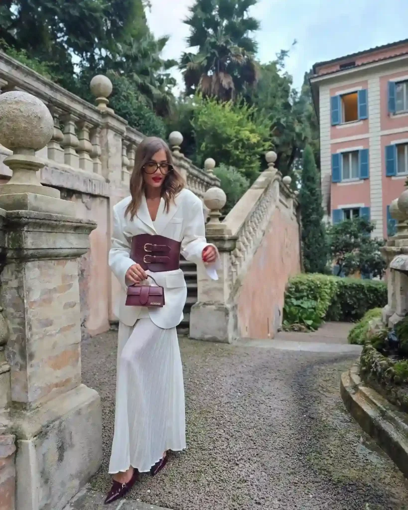 A stylish woman in a white dress and brown belt poses on a garden path with lush greenery and elegant architecture.