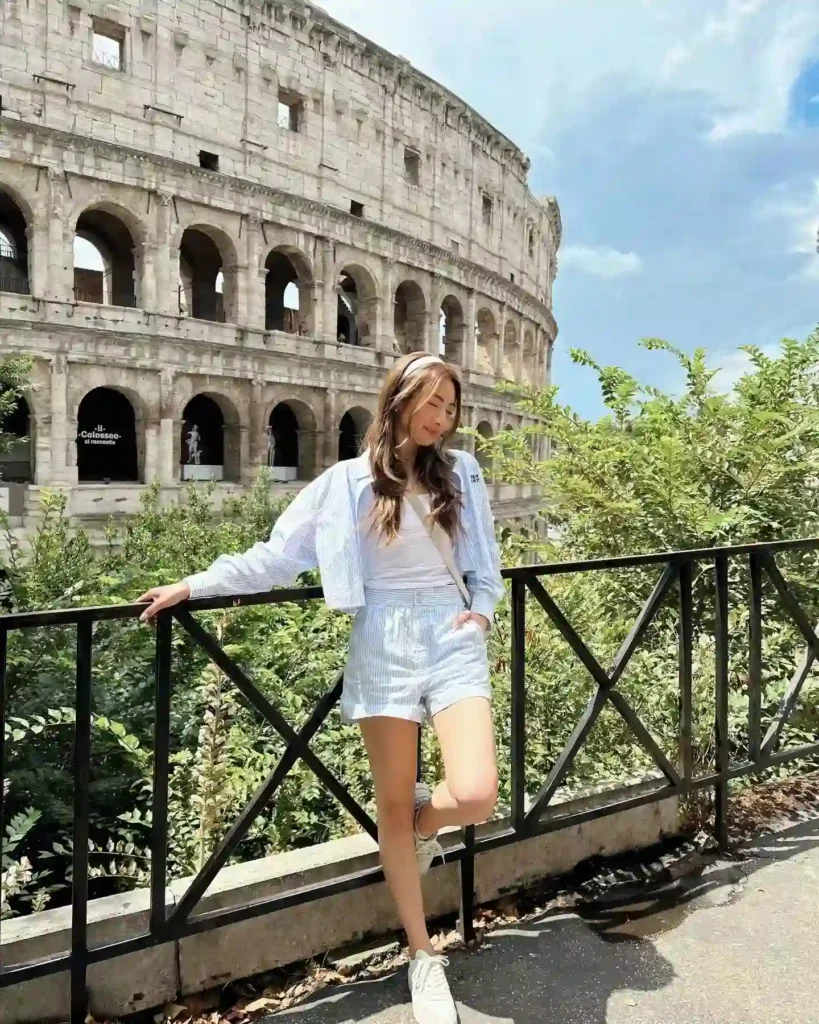 Young woman in a striped outfit posing near the Colosseum under a bright sky, surrounded by greenery.
