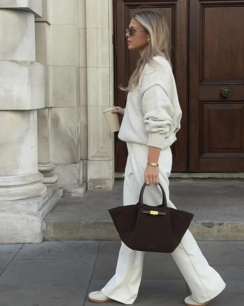 Fashionable woman walking while holding a coffee cup and a chic brown tote bag, wearing stylish sunglasses and a cozy sweater.