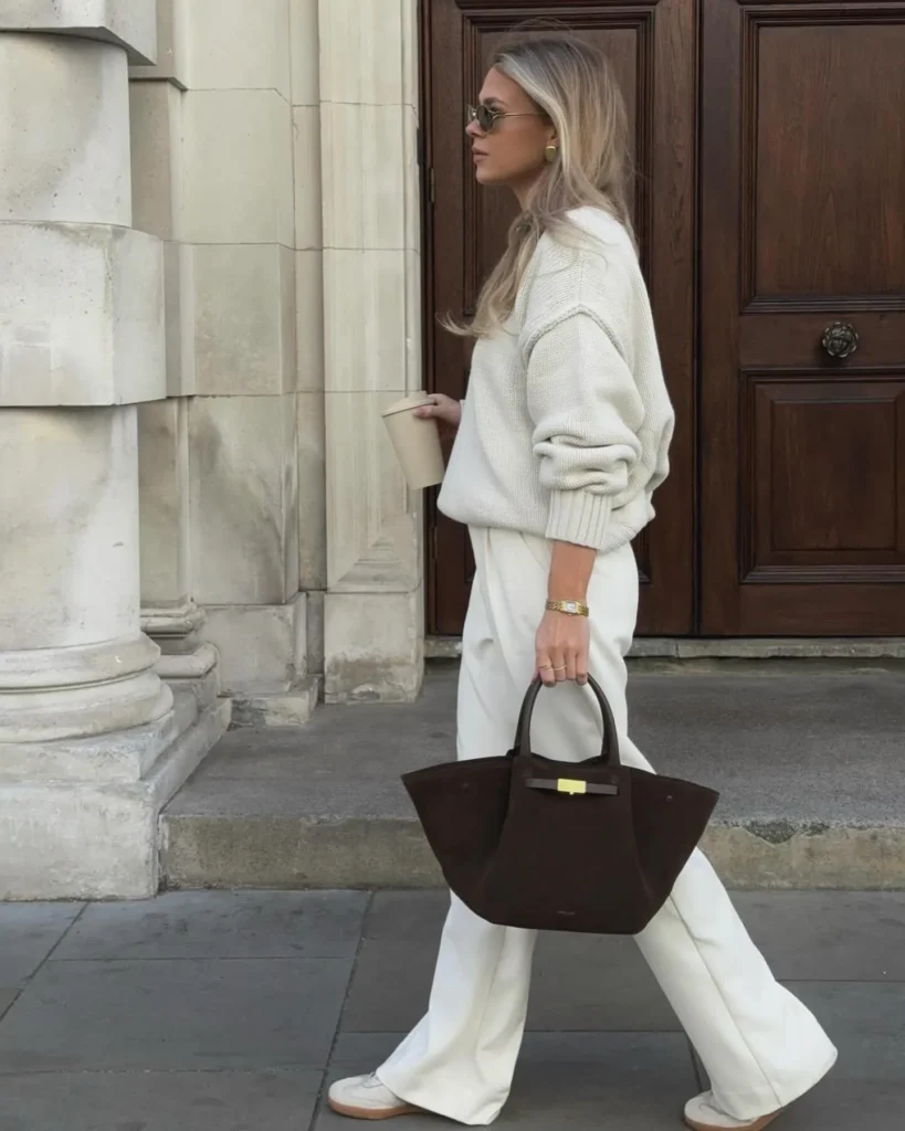 Stylish woman with long hair in sunglasses, wearing a cozy sweater and white pants, holding a coffee and handbag.