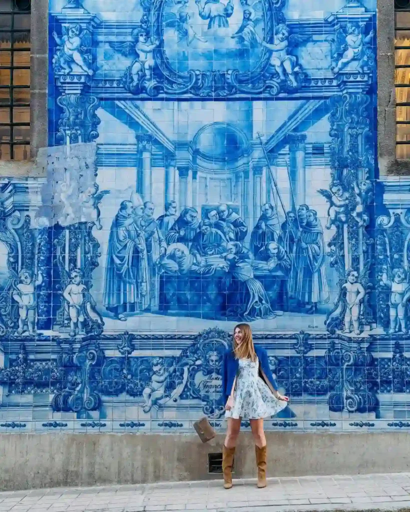 Woman in a floral dress poses in front of a vibrant blue tile mural, showcasing intricate art and historical scenes.
