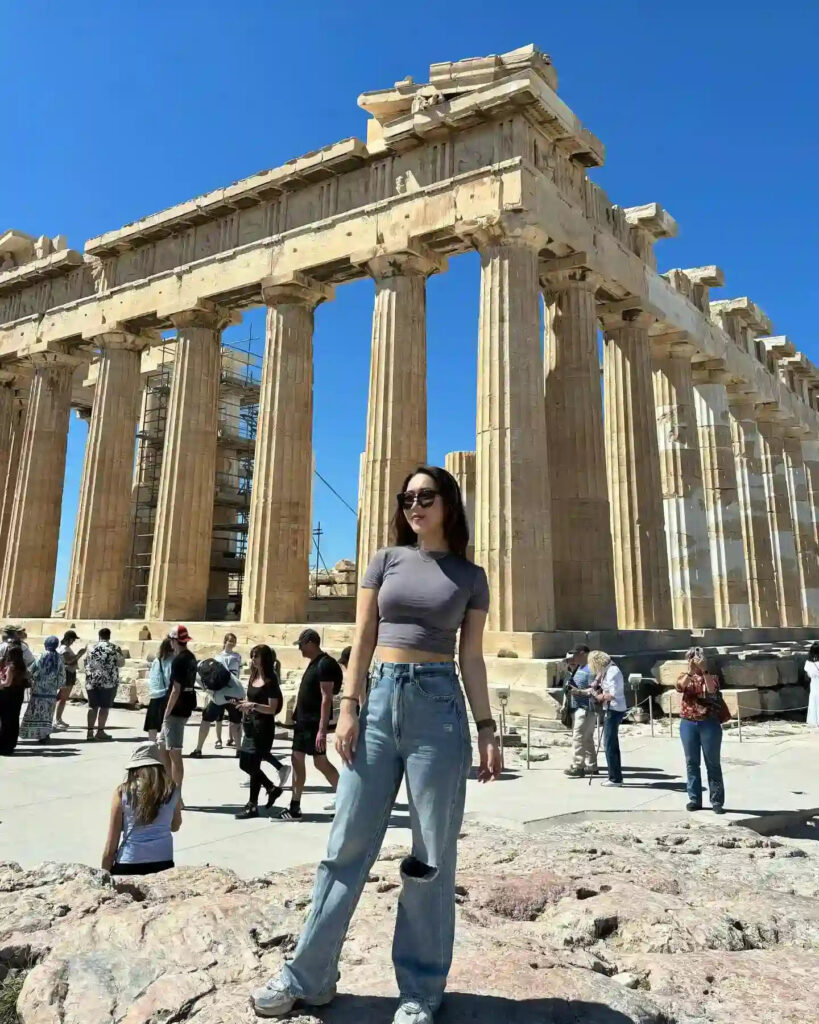 Young woman posing in front of the Parthenon in Athens, surrounded by tourists under a clear blue sky.