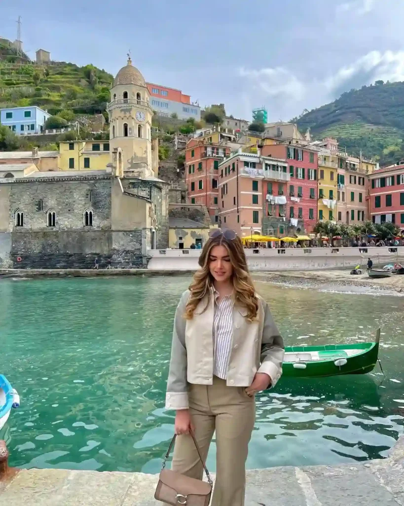 A young woman in stylish attire stands by a scenic harbor, surrounded by colorful buildings and green hills.