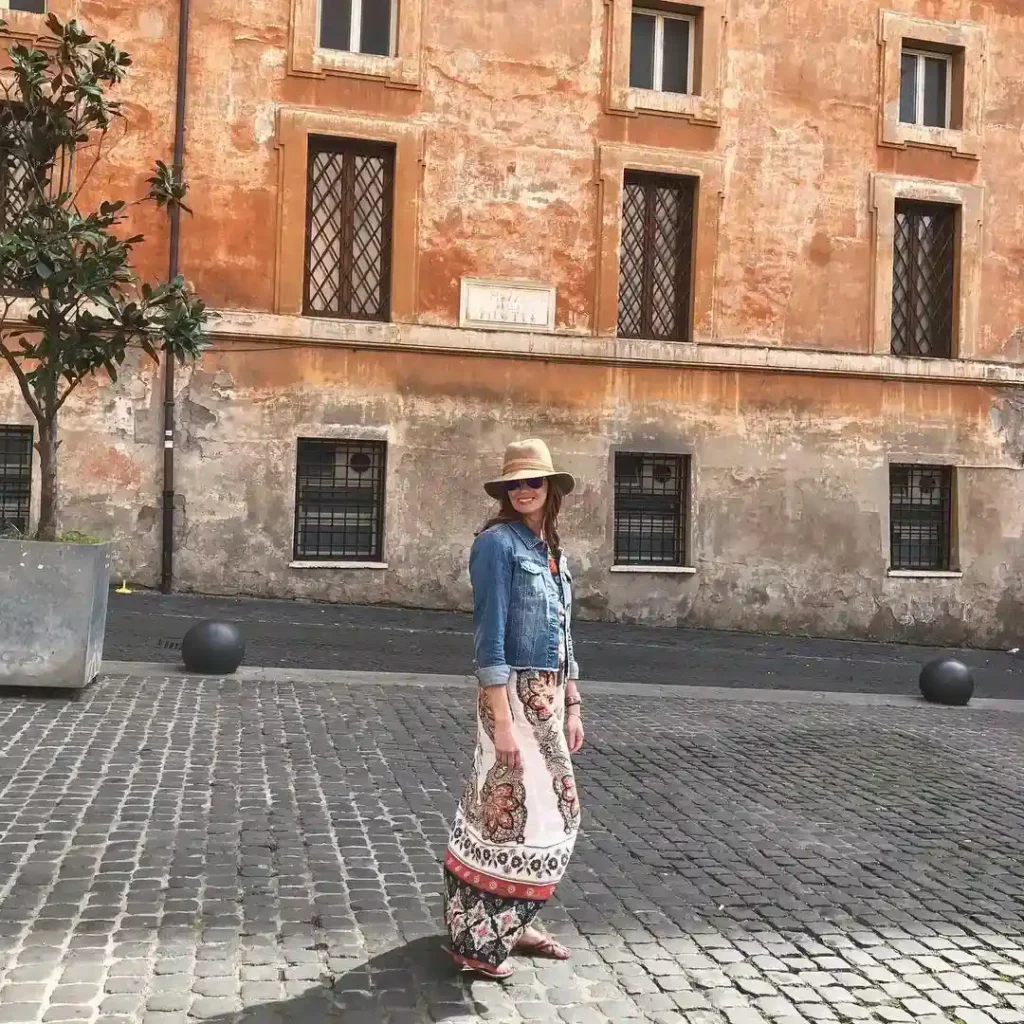A woman in a stylish outfit and hat stands in a charming cobblestone street near an old building.