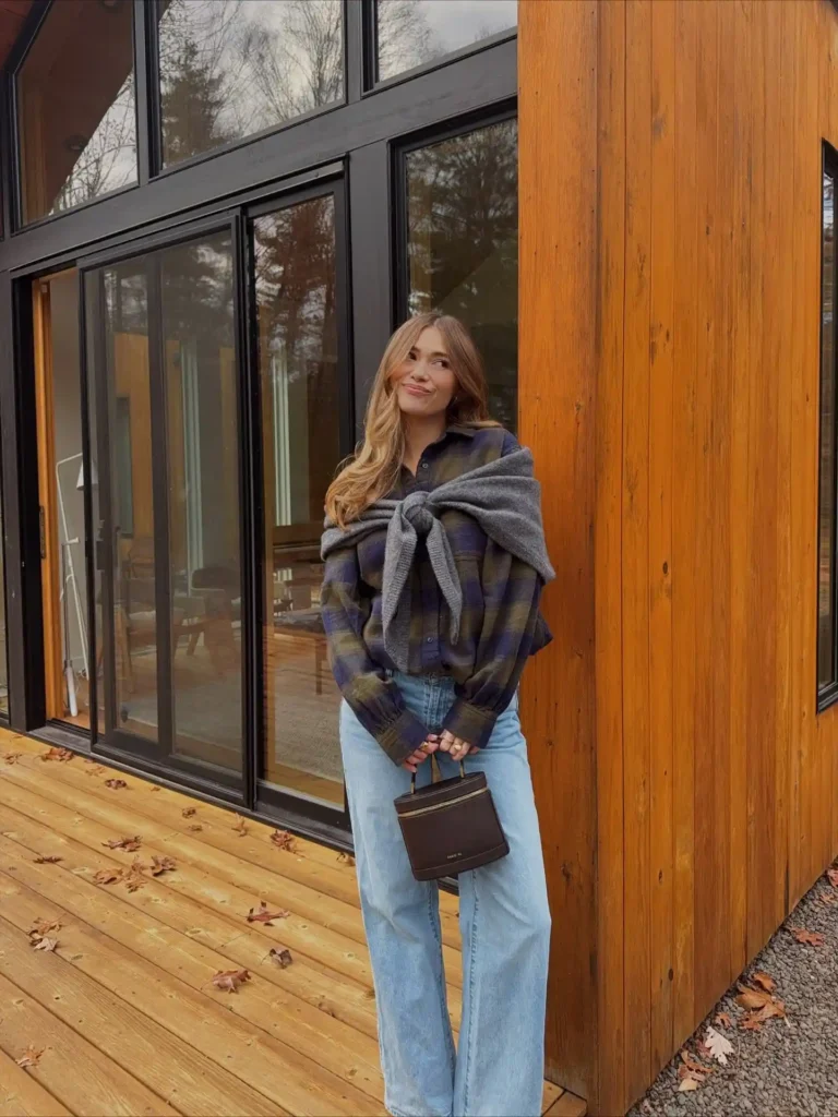 A woman in a plaid shirt stands outside a modern wooden house, smiling while holding a small handbag.