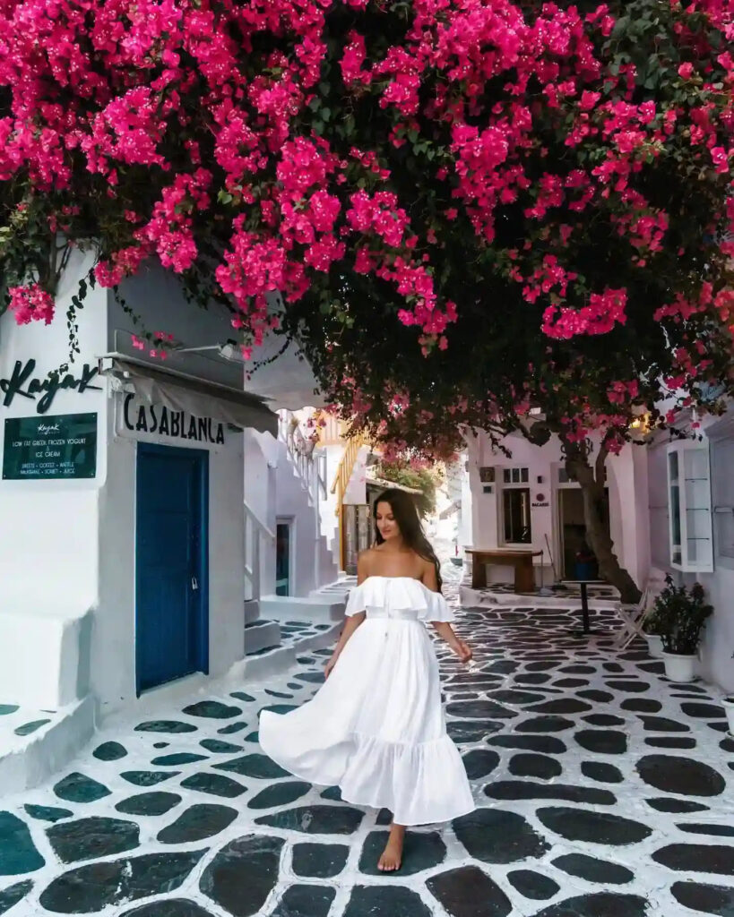 A woman in a white dress strolls through a charming, flower-covered street in a Mediterranean village.