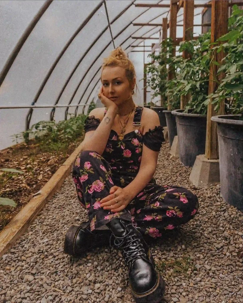 Young woman in floral jumpsuit sitting in a greenhouse surrounded by potted plants, creating a serene, natural atmosphere.