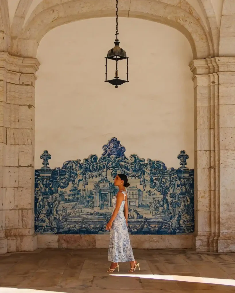 Woman in a floral dress stands elegantly in a historic archway with blue tile artwork and vintage lantern.