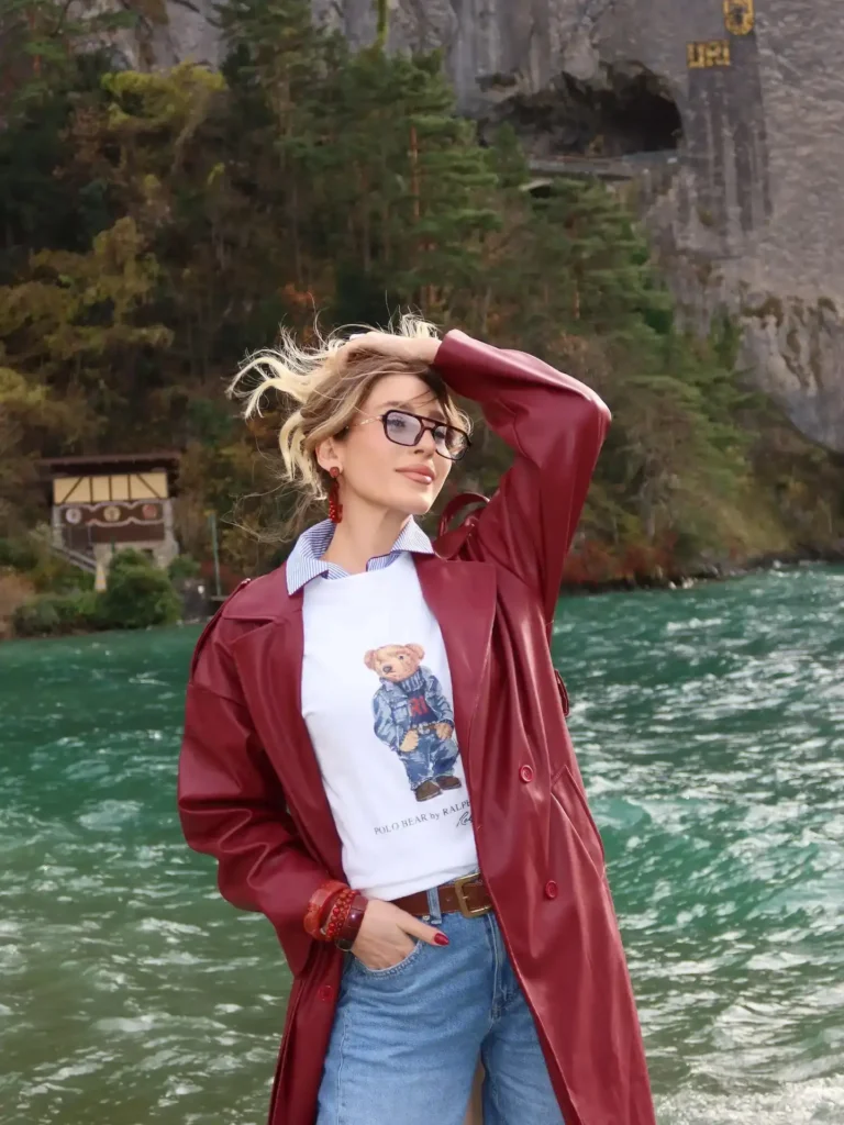 Woman in a red coat poses confidently by a river, wearing a bear graphic tee and glasses, with nature in the background.
