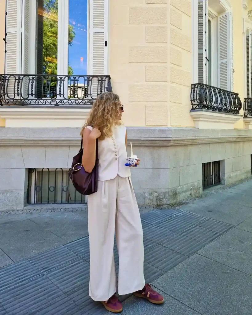 A stylish woman in a beige outfit and sunglasses stands on the street, holding a drink near elegant architecture.