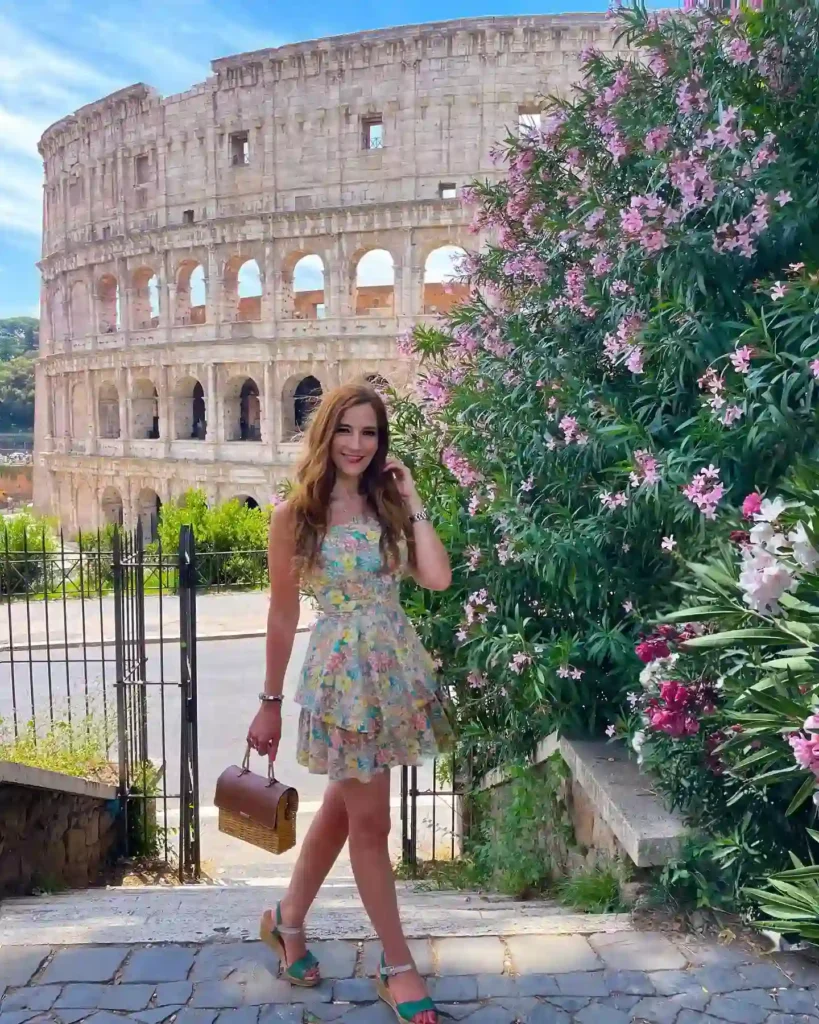 Young woman in a floral dress poses by the Colosseum, surrounded by blooming flowers on a sunny day in Rome.