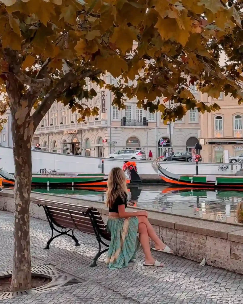 A woman sits by a scenic waterfront, adorned with golden leaves and colorful boats, enjoying a peaceful moment.