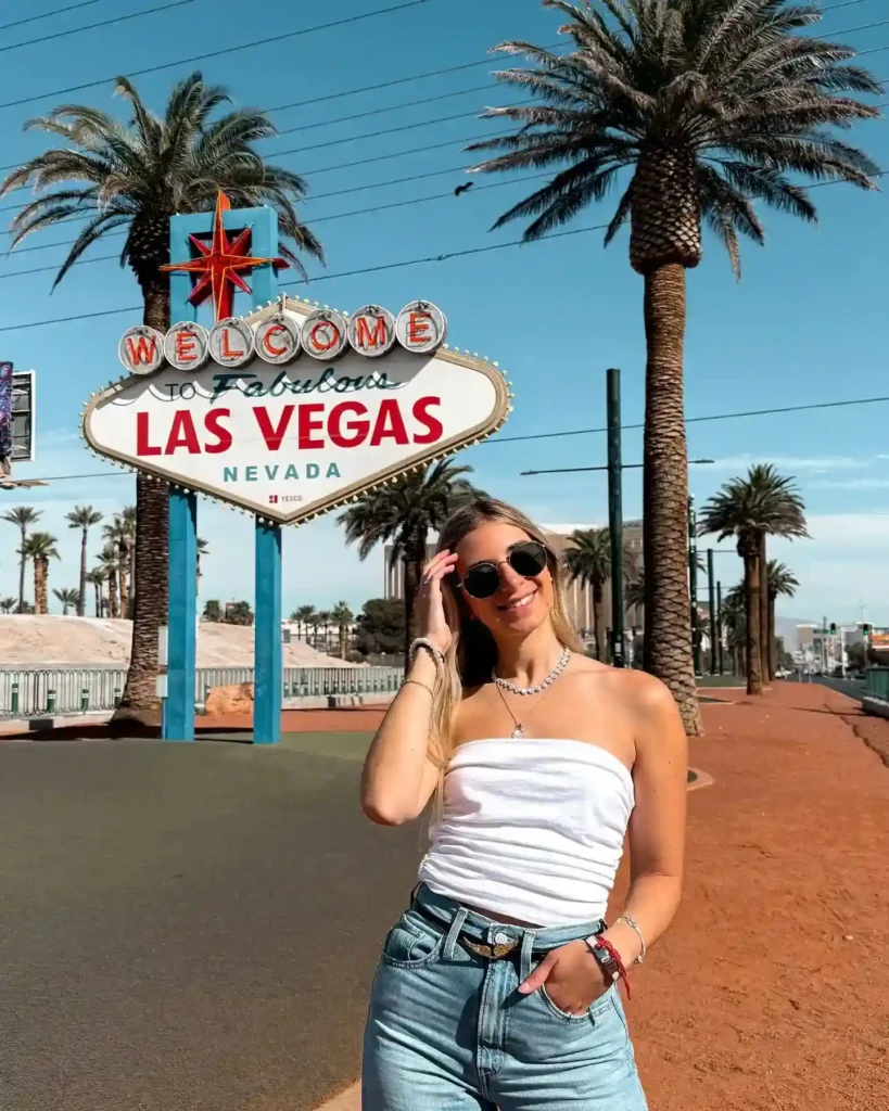 Young woman poses in front of the iconic Welcome to Fabulous Las Vegas sign, surrounded by palm trees and clear skies.