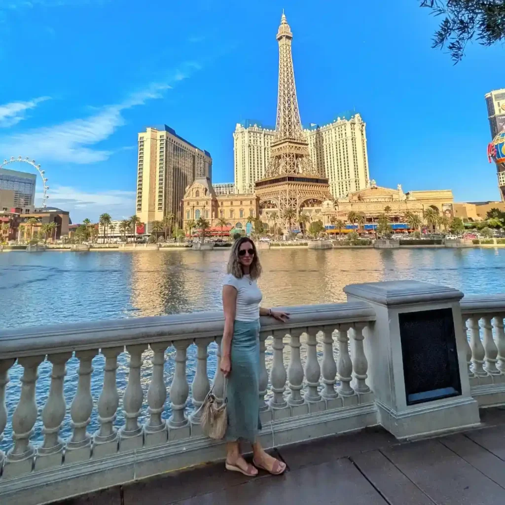 Woman posing by the water near the Eiffel Tower replica in Las Vegas, with blue skies and palm trees in the background.