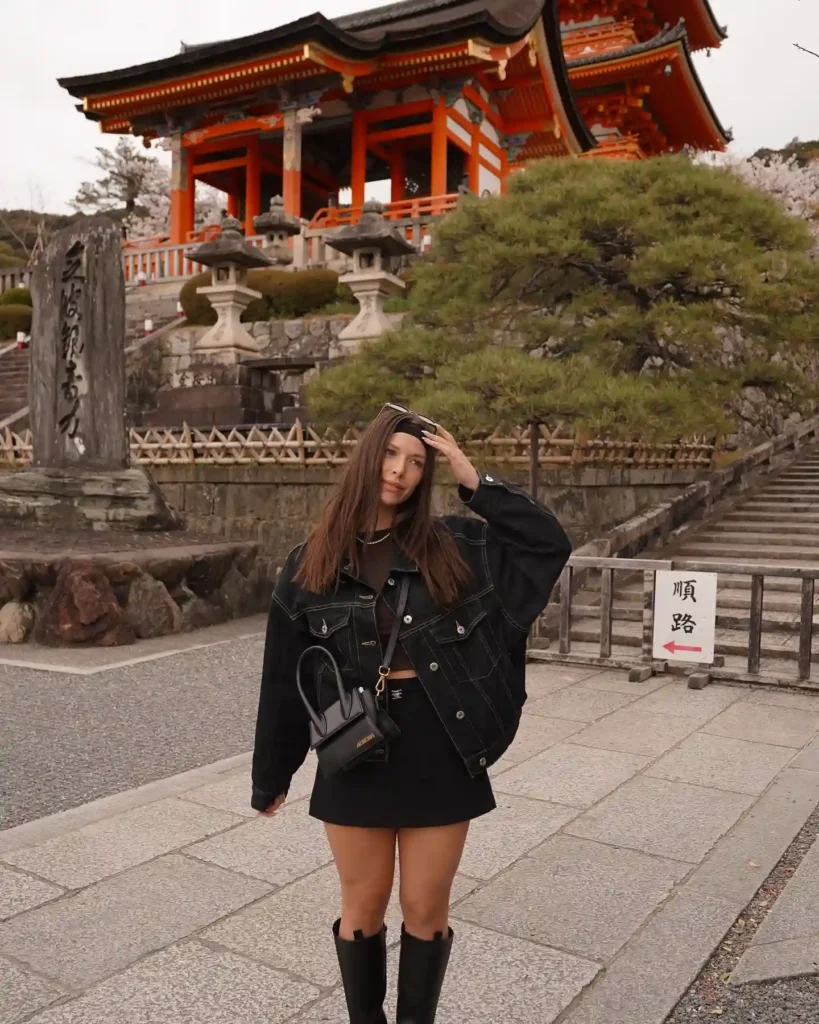 Young woman in a black outfit poses in front of a traditional Japanese temple with cherry blossoms in a serene setting.