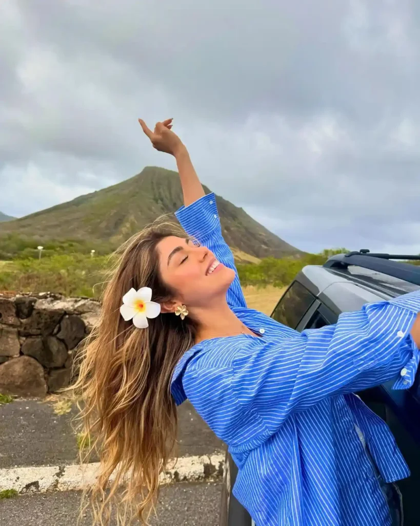 Girl with long hair and a flower in her hair joyfully poses beside a car, with a mountain backdrop under a cloudy sky.