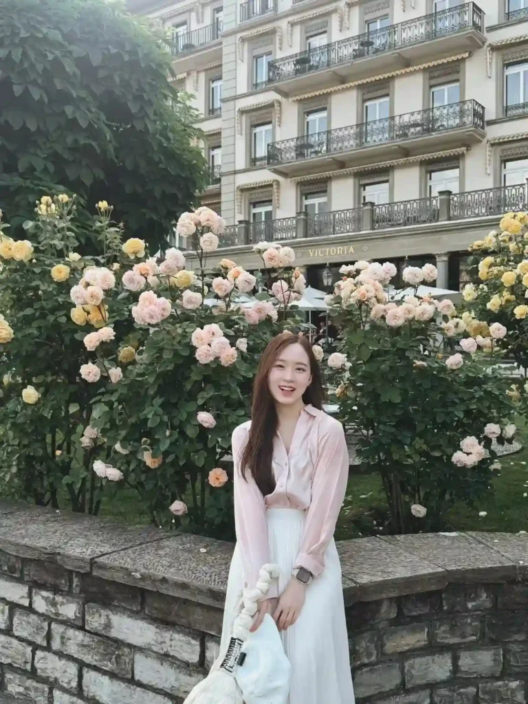 Young woman in a pink blouse and white skirt smiles by a rose garden near a historic building.