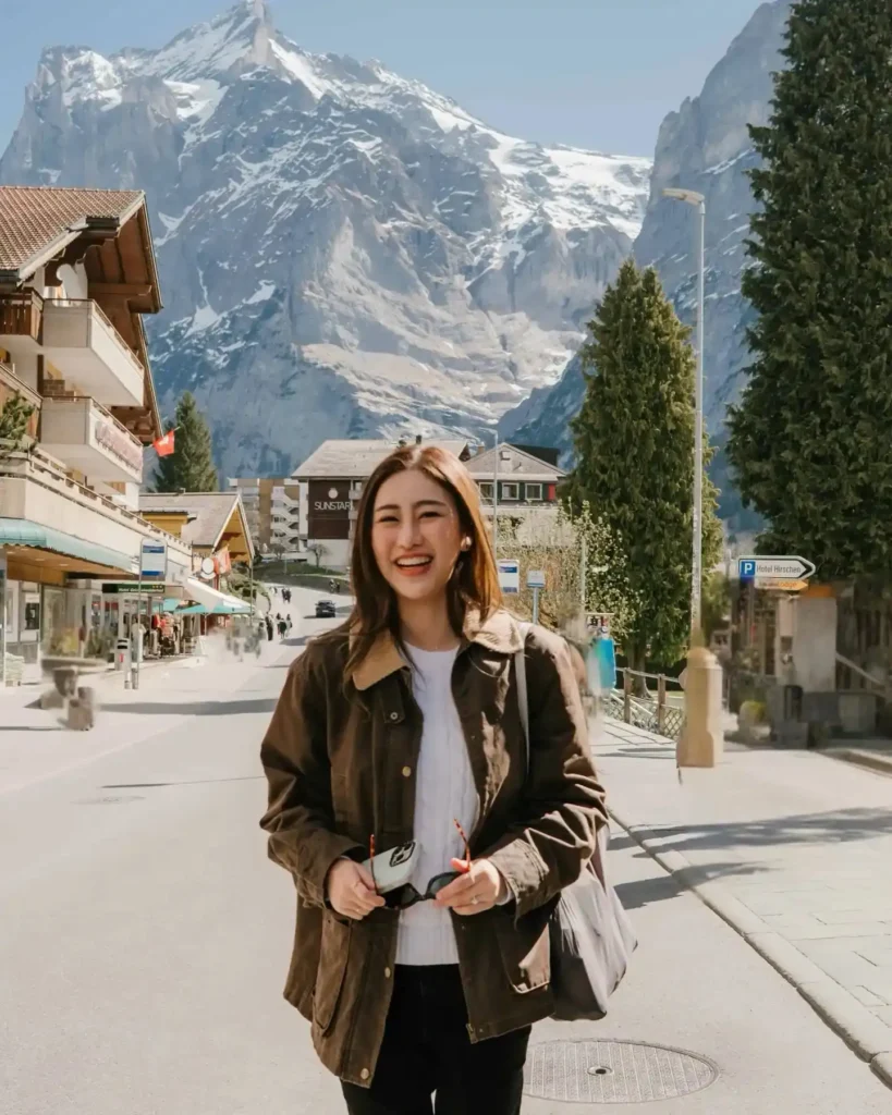 A smiling woman stands on a street in a charming town, surrounded by mountains and shops on a sunny day.