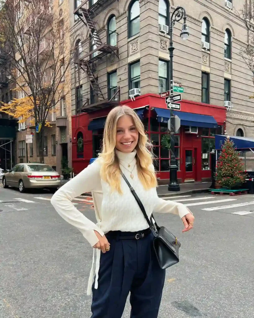Young woman smiling on a city street, wearing a white sweater and black pants, with festive decorations nearby.