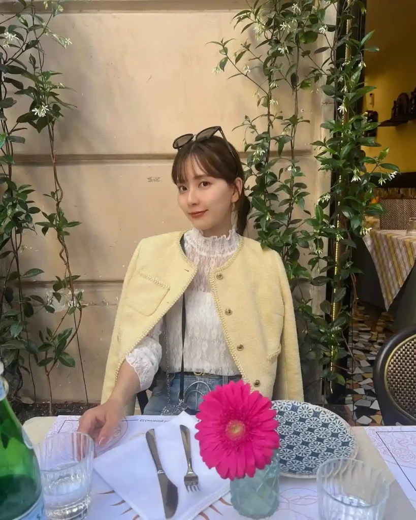 A stylish woman in a yellow jacket poses at a beautifully set table with pink flowers and greenery in a café.