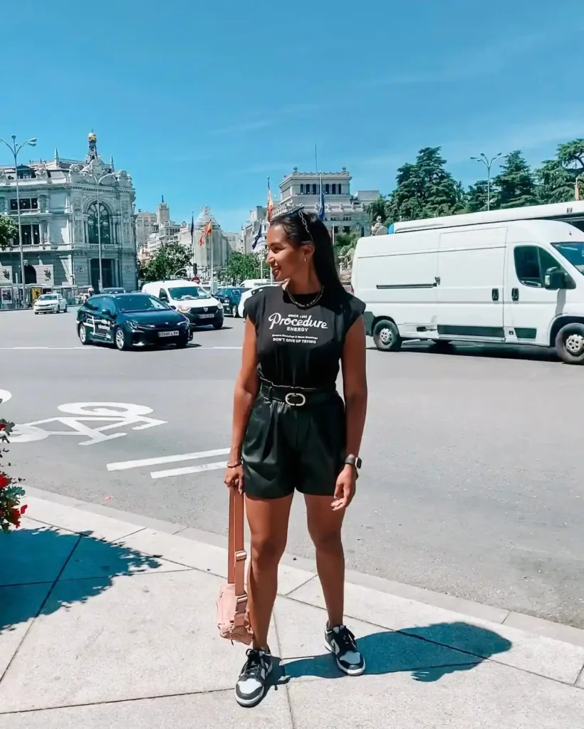 Woman in stylish black outfit and sneakers stands on a city street with cars and buildings in the background on a sunny day.