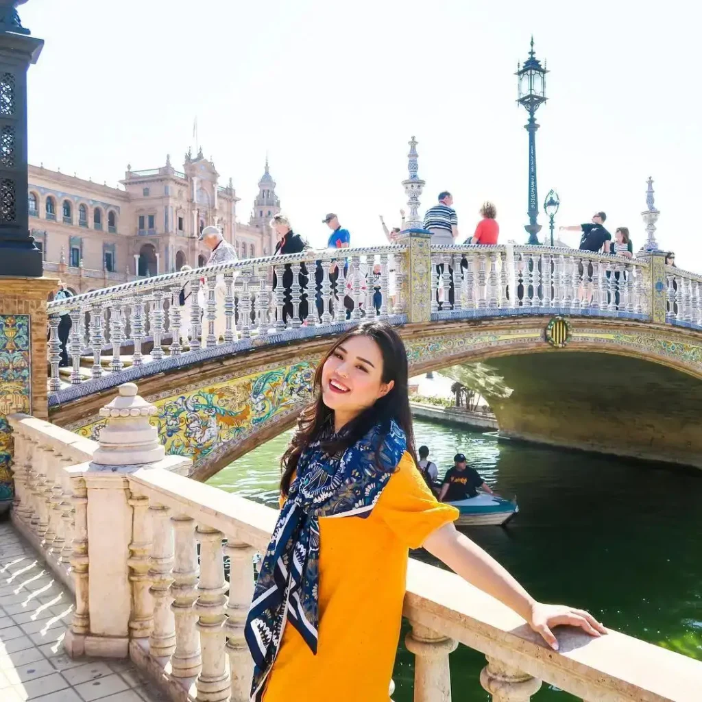 Smiling woman in a yellow dress poses by a colorful bridge, with a scenic canal and historic buildings in the background.