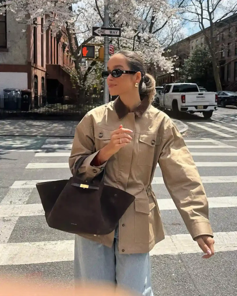 A stylish woman in a beige jacket and sunglasses walks across a city street lined with cherry blossom trees.