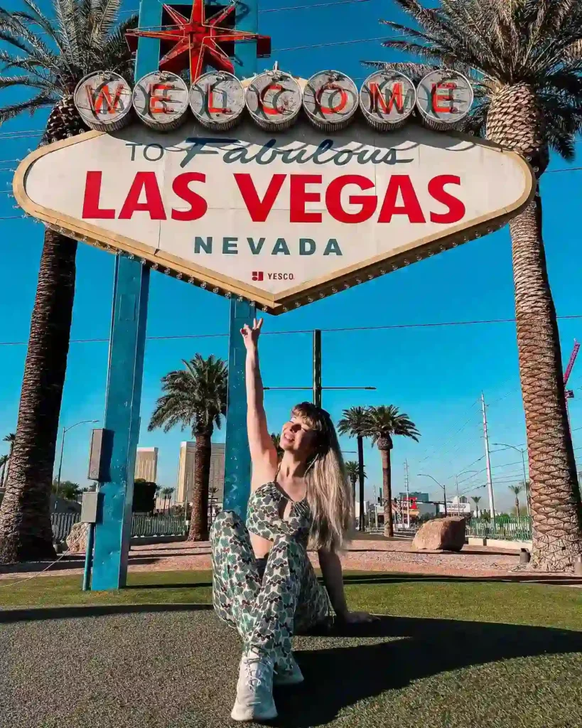 Woman celebrating in front of the iconic Welcome to Las Vegas sign, surrounded by palm trees and blue skies.