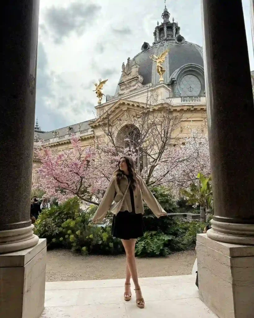 Woman in a stylish outfit poses in front of a beautiful historic building surrounded by blooming cherry trees.