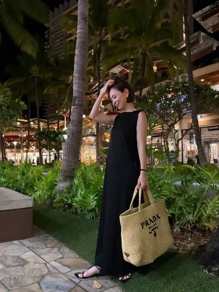 Woman in a black dress holding a Prada bag, enjoying an evening in a tropical, well-lit outdoor setting.