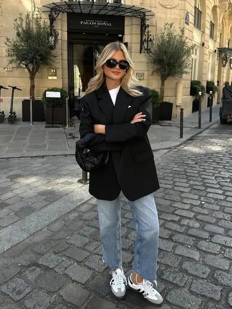 Fashionable woman in a black blazer and sunglasses poses stylishly on a cobblestone street near Palais Royal.