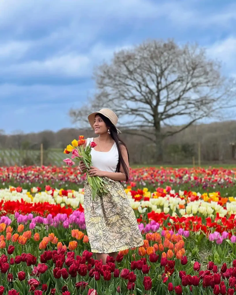 Woman in a hat wearing a floral skirt holds colorful tulips in a vibrant tulip field under a blue sky.