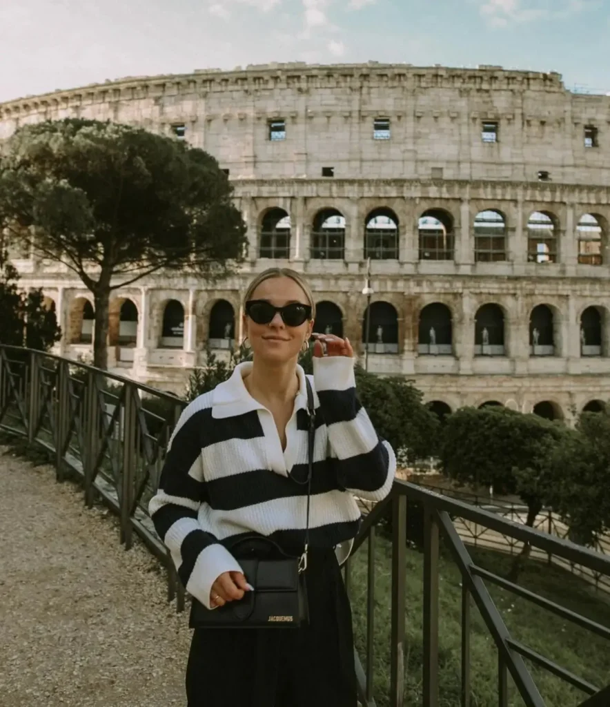A stylish woman in sunglasses stands near the Colosseum in Rome, showcasing a casual outfit with a chic bag.