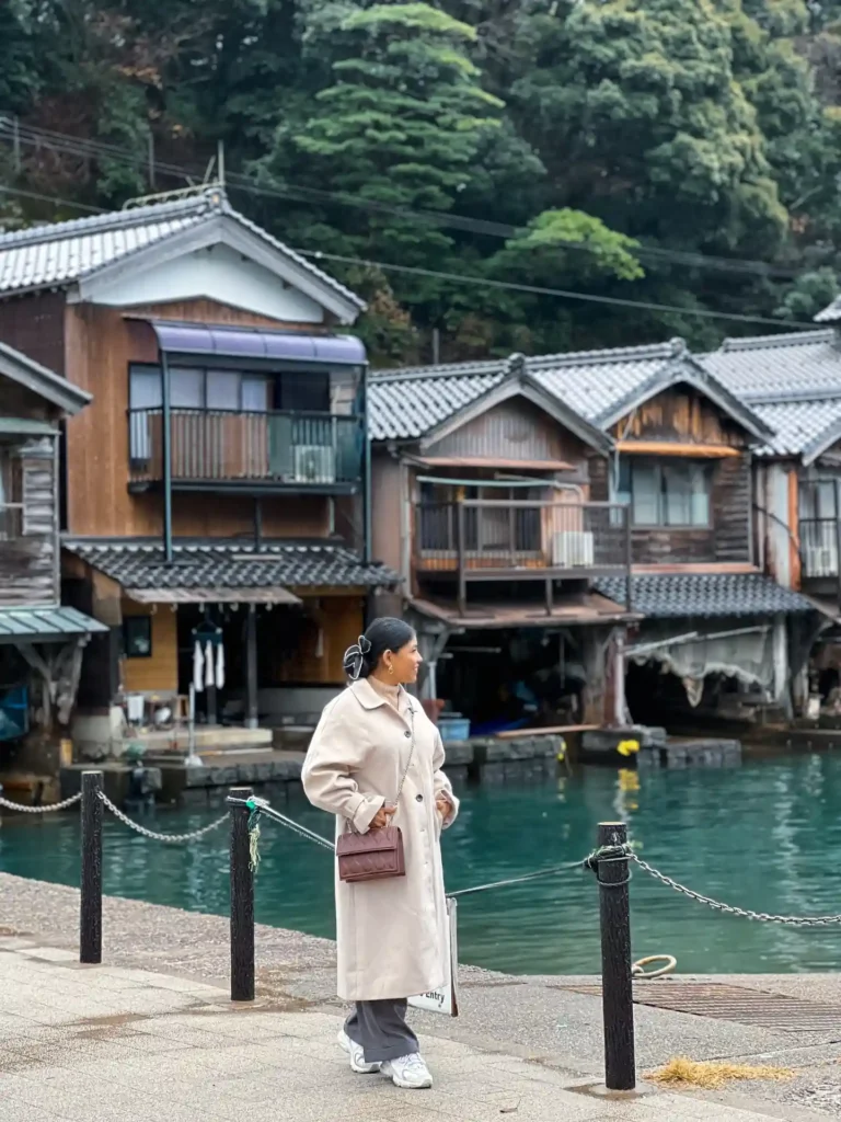 A woman in a beige coat strolls by traditional wooden houses along a serene waterfront surrounded by lush greenery.
