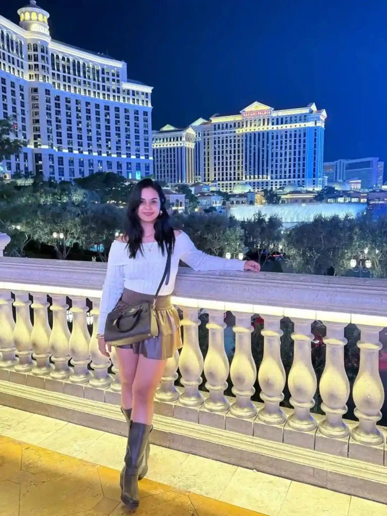 Smiling woman in a stylish outfit poses at a lit balcony with a view of luxury hotels in Las Vegas at night.