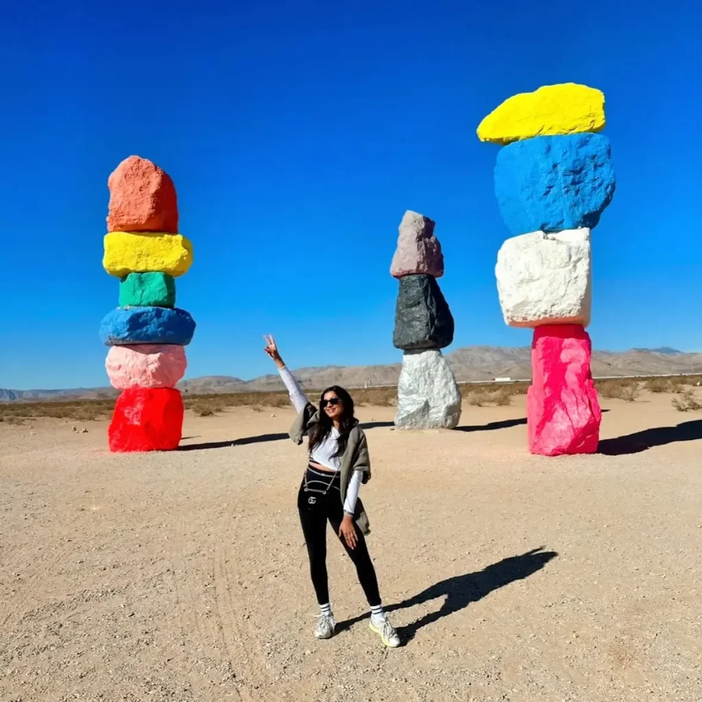Person posing joyfully in front of colorful rock formations against a clear blue sky at a desert location.