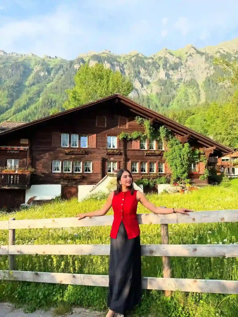 Young woman in red vest poses near a wooden house amid lush green mountains and vibrant nature.