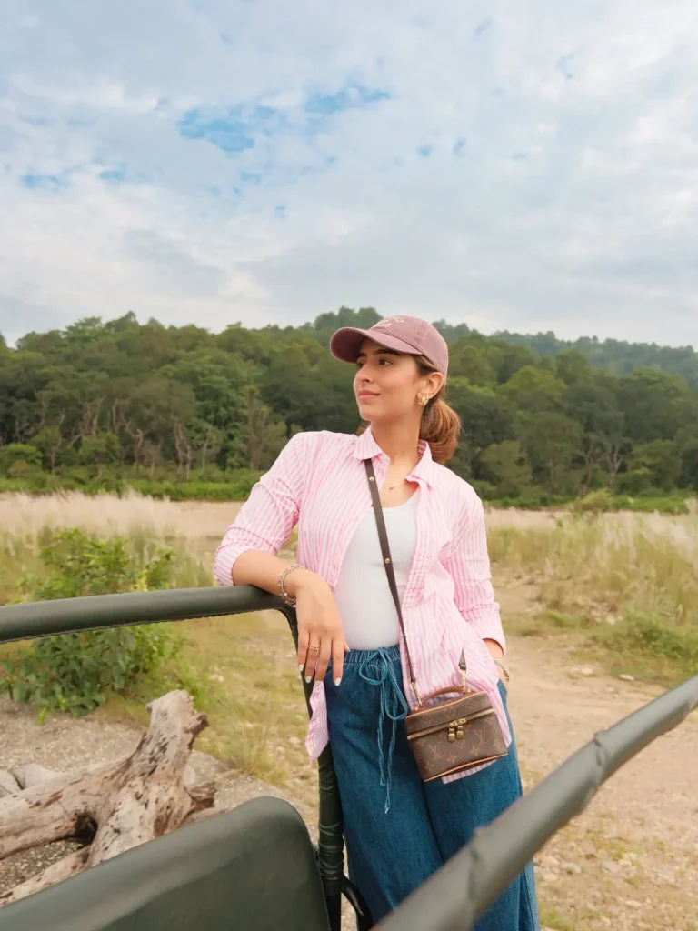 Woman in a pink striped shirt and denim pants poses stylishly outdoors, surrounded by greenery and a cloudy sky.