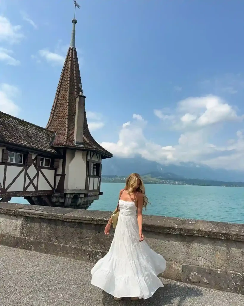Woman in a flowing white dress twirls near a scenic lakeside with a charming house and mountains in the background.