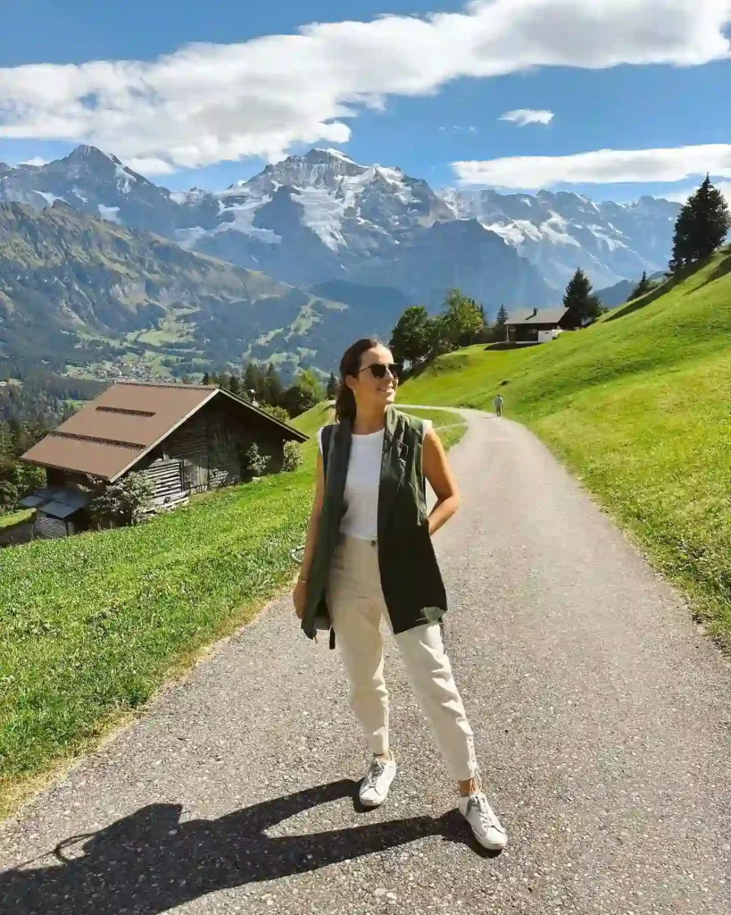 Young woman in casual attire standing on a scenic path with mountains and green hills in the background.
