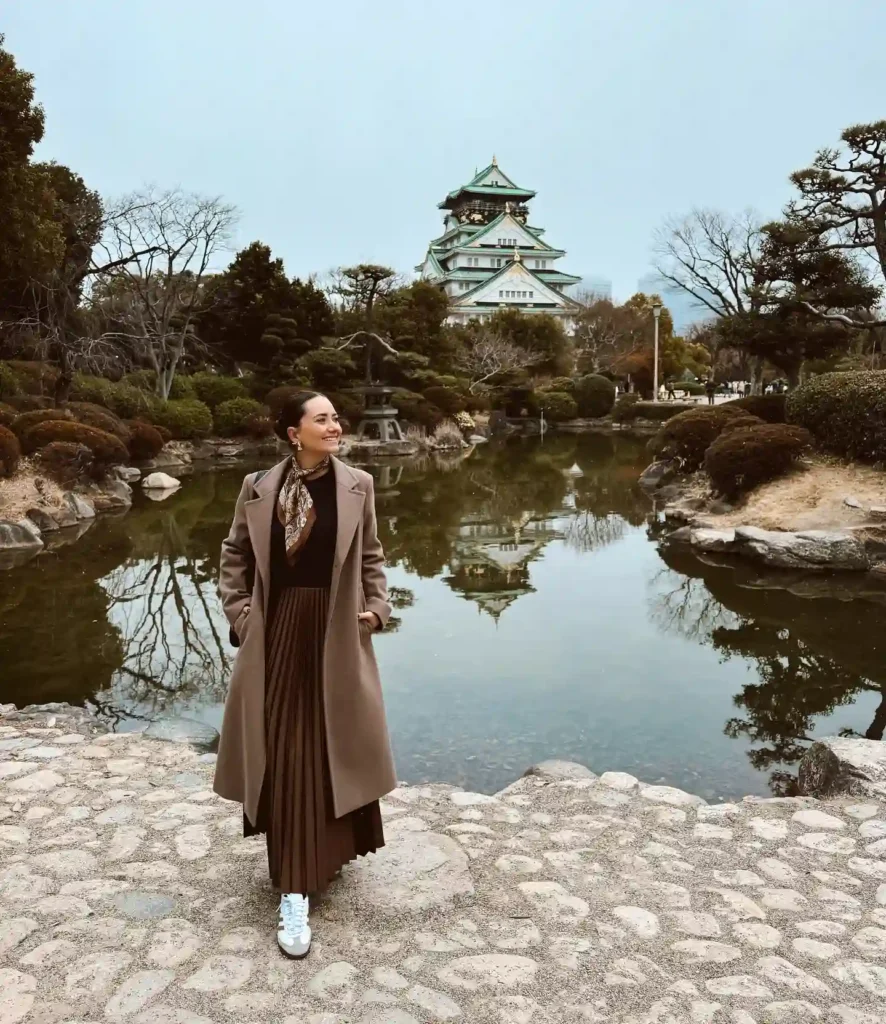 Woman in a brown coat stands by a serene pond with Osaka Castle in the background, surrounded by lush greenery.