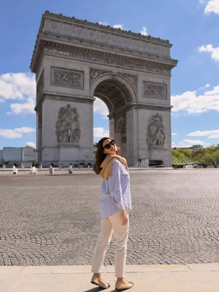 Woman posing in front of the Arc de Triomphe in Paris on a sunny day, wearing sunglasses and a stylish outfit.