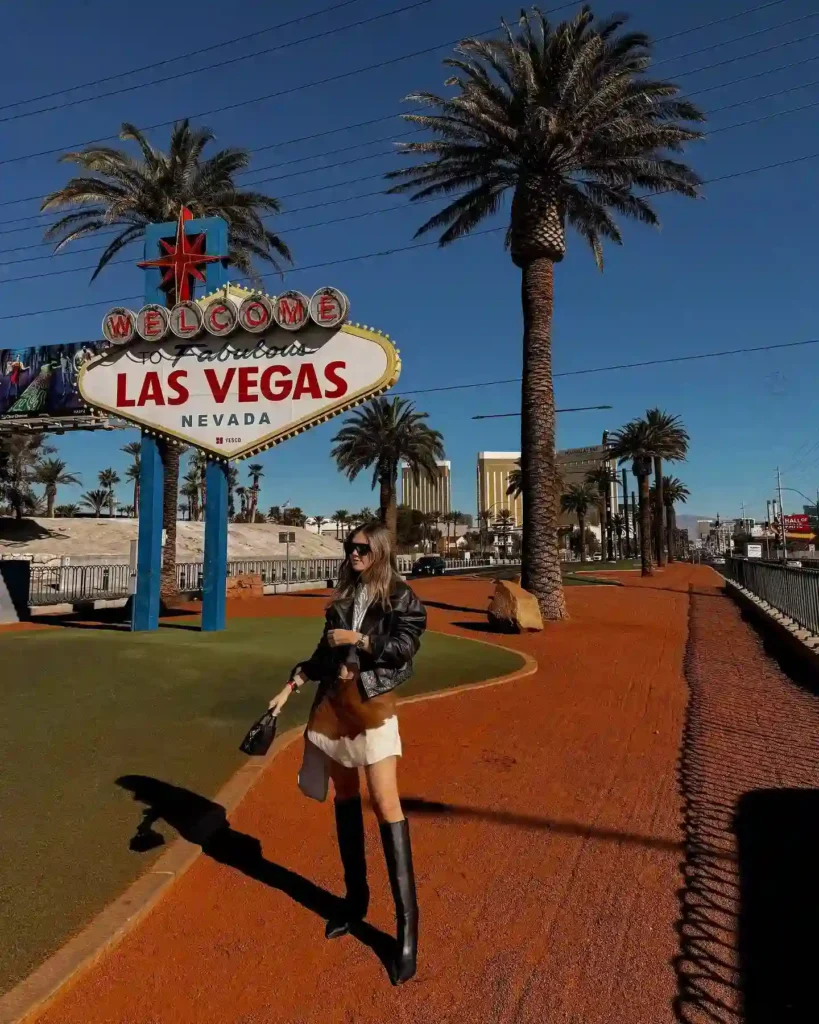 Fashionable woman poses near the iconic Welcome to Las Vegas sign, surrounded by palm trees and a vibrant blue sky.