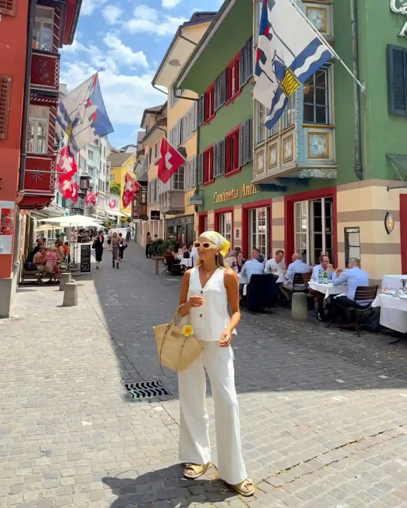 A woman in a white outfit stands in a vibrant Swiss street lined with flags and outdoor dining. Sunny day in Zurich.