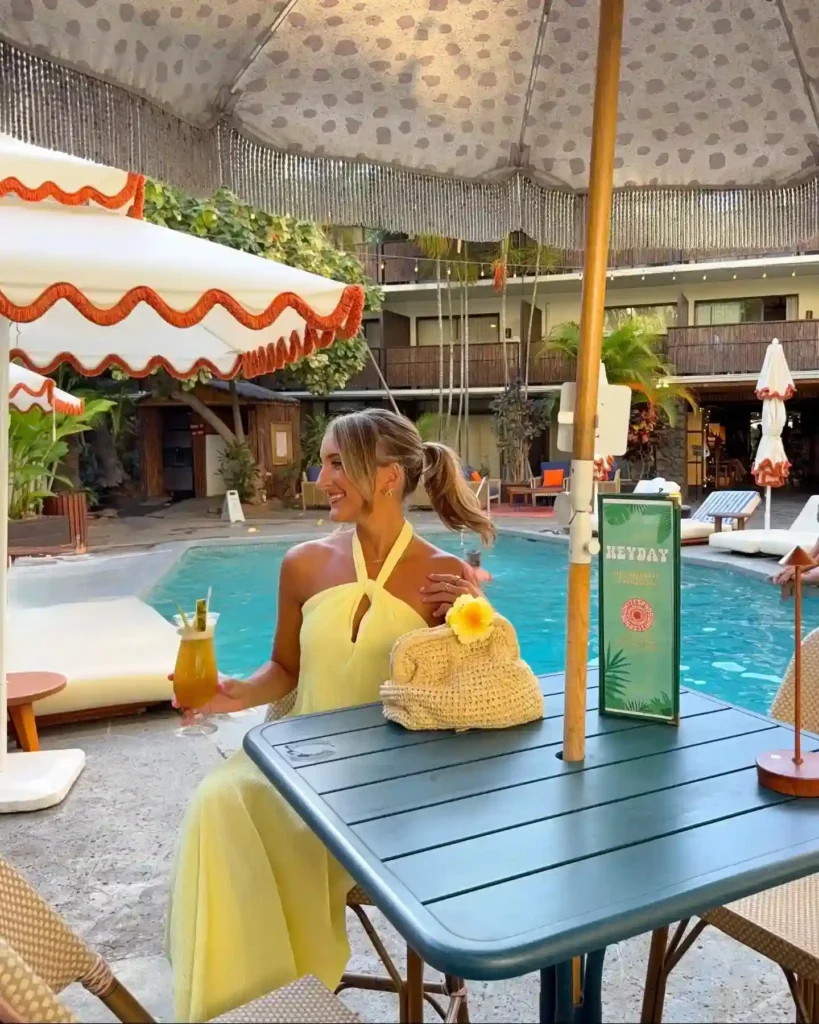 Woman in a yellow dress enjoying a cocktail by the poolside, surrounded by lush greenery and vibrant decor.