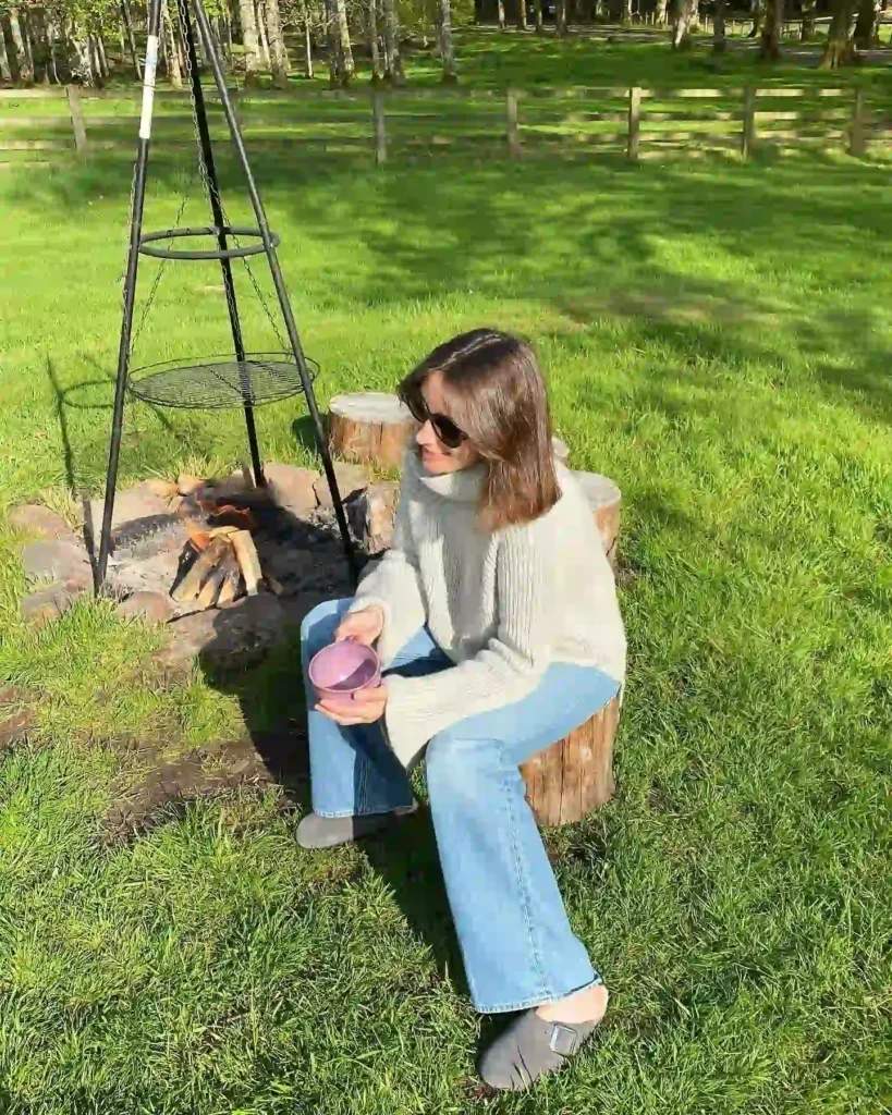 Woman in a cozy sweater sits on a log by a campfire, enjoying a drink in a green outdoor setting.