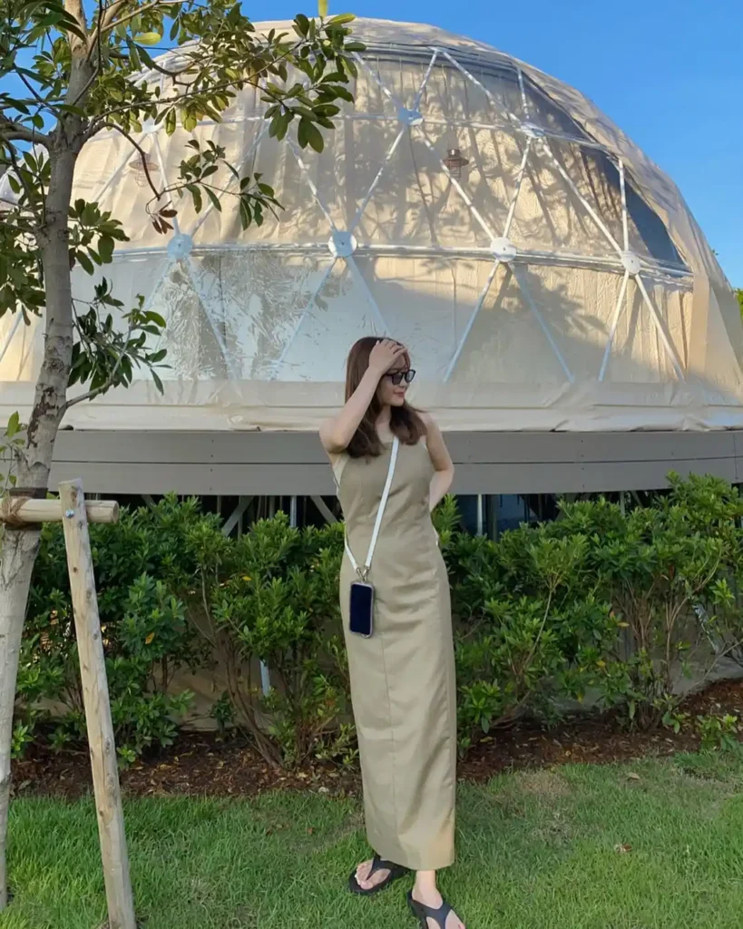 Woman in a tan dress poses in front of a geodesic dome structure, surrounded by greenery and a clear blue sky.