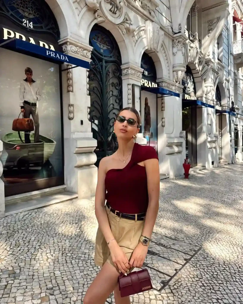 Stylish woman in a one-shoulder top poses outside Prada storefront in a vibrant city setting.